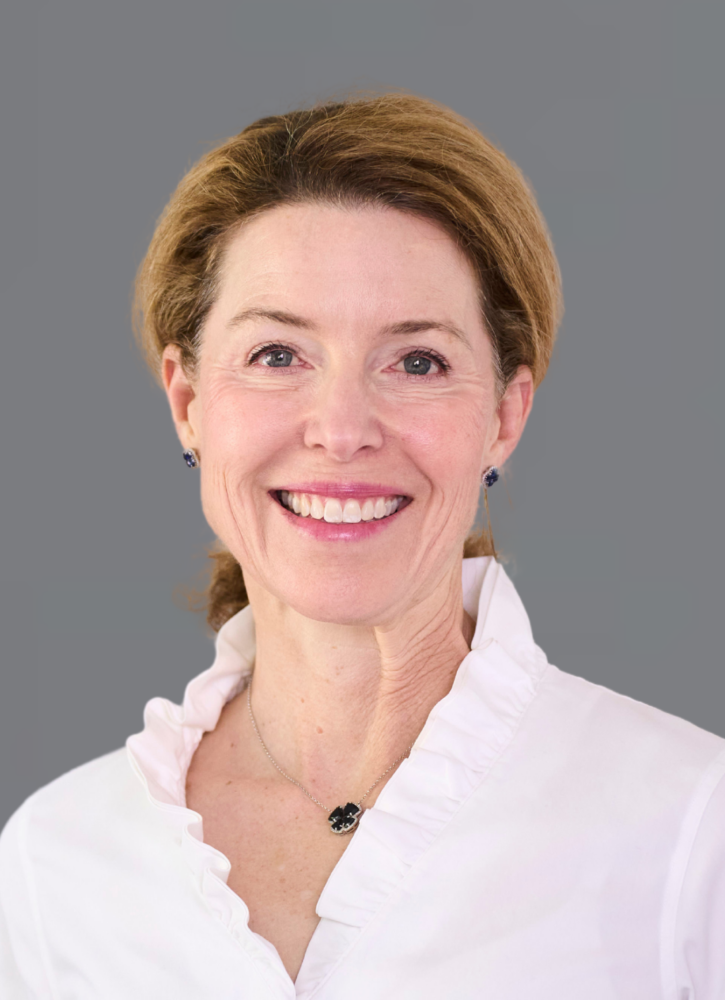 Smiling person with light brown hair wearing a white blouse and necklace, against a gray background.